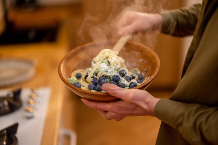 Foto de Hombre mezclando sabrosa pasta caliente con arándanos y espinacas en un tazón de madera, primer plano - Imagen libre de derechos