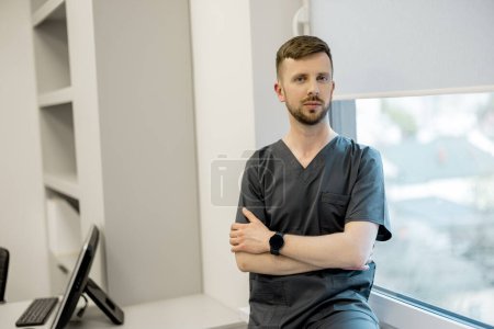 Téléchargez les photos : Portrait de jeune médecin ou praticien en uniforme assis sur le rebord de la fenêtre au bureau - en image libre de droit