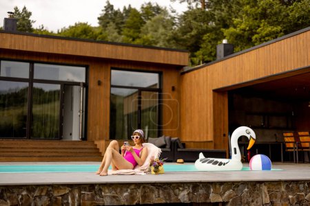 Téléchargez les photos : Jeune femme utilisant le téléphone tout en bronzant près de la maison avec une piscine, passer des vacances d'été - en image libre de droit