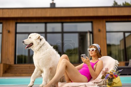 Téléchargez les photos : Femme bronzant tout en se relaxant avec son chien près de la maison avec une piscine. Amitié avec les animaux domestiques et concept de vacances d'été - en image libre de droit
