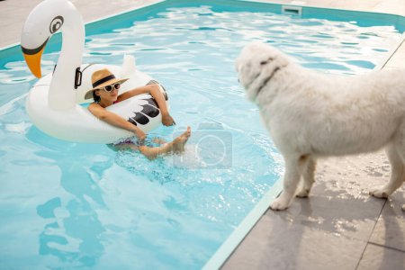 Téléchargez les photos : Femme nage sur le flamant rose gonflable et joue avec son chien blanc mignon, se reposant et passant l'heure d'été avec l'animal de compagnie à la piscine - en image libre de droit