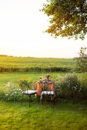 Foto de Beautiful green lawn with flowers and dining table for two during the sunset. Romantic picnic on nature - Imagen libre de derechos
