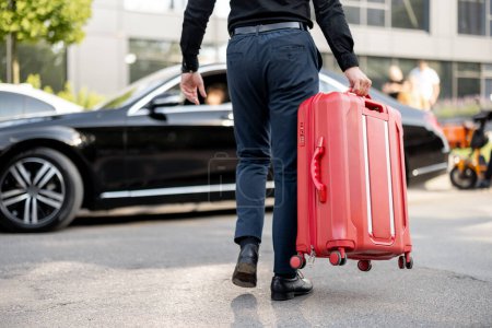Téléchargez les photos : L'homme porte une valise rouge jusqu'à une voiture, vue de dessous. Concept de service de chauffeur et voyages d'affaires - en image libre de droit