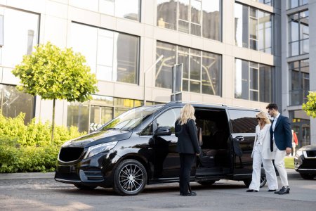 Photo for Female chauffeur waits a business people to let them in a minivan taxi, keeping door open. Concept of business trips and transportation service - Royalty Free Image