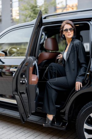 Photo for Portrait of an elegant business lady in black getting out of a luxury SUV taxi. Concept of business trips and transportation - Royalty Free Image