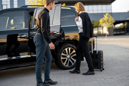 Téléchargez les photos : Chauffeur aide l'homme d'affaires avec une valise pour monter dans la voiture, ouvrant une porte d'un taxi noir de luxe. Homme en voyage d'affaires - en image libre de droit