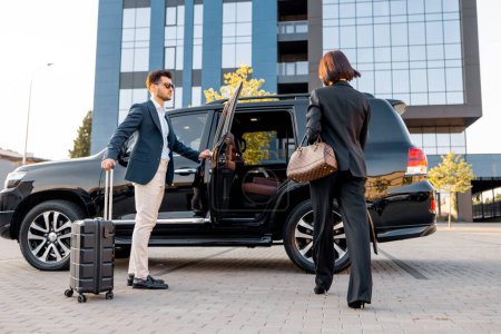 Photo for Elegant business woman walks to the luxury car, man with a suitcase opens vehicle door, letting lady in. Concept of business trip and travel - Royalty Free Image