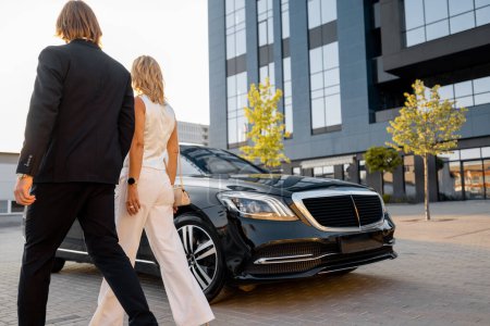 Photo for Businessman and businesswoman walk to a luxury black car near hotel or office building on sunset. Concept of transportation and business travel - Royalty Free Image