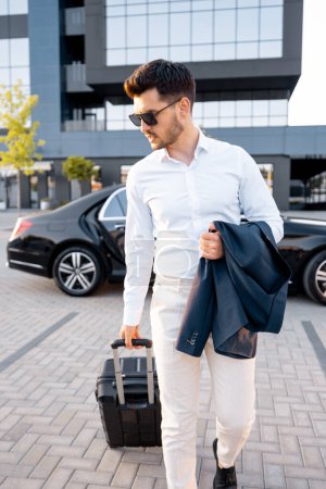 Téléchargez les photos : Élégant homme d'affaires marche avec une valise, portant une veste devant la voiture près du bâtiment moderne au coucher du soleil - en image libre de droit