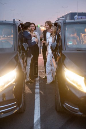 Téléchargez les photos : Groupe de partenaires d'affaires s'amusent ensemble, boire de l'alcool tout en célébrant entre les voitures sur le parking la nuit - en image libre de droit