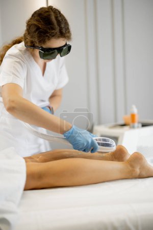 Téléchargez les photos : Jeune travailleur d'un salon de beauté lors de la procédure d'épilation sur les jambes d'une femme. Concept de dépilation et procédures beauté - en image libre de droit