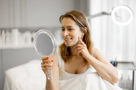 Téléchargez les photos : Femme heureuse regarde dans le miroir sur son visage après les procédures de beauté tout en étant assis au salon. Concept de beauté et de soin du visage - en image libre de droit