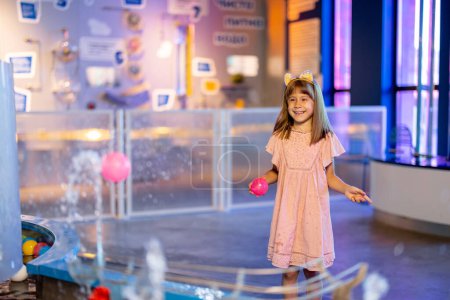 Téléchargez les photos : Petite fille joue avec une balle sur une vapeur d'eau, apprenant des phénomènes physiques d'une manière intéressante, s'amusant dans un musée des sciences avec des modèles interactifs - en image libre de droit