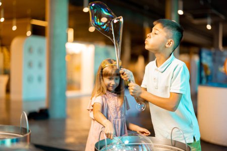 Foto de Niña y niño hacen burbujas de jabón, mientras juegan juntos y se divierten en un museo de ciencias. Concepto de entretenimiento y aprendizaje infantil - Imagen libre de derechos