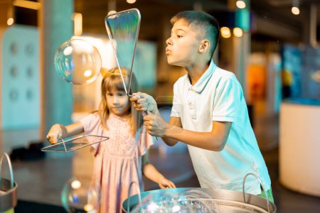 Foto de Niña y niño hacen burbujas de jabón, mientras juegan juntos y se divierten en un museo de ciencias. Concepto de entretenimiento y aprendizaje infantil - Imagen libre de derechos