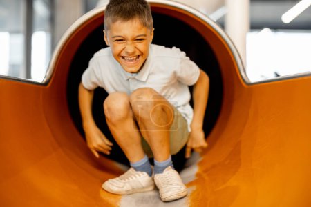 Téléchargez les photos : Garçon heureux descend un tube, tout en jouant dans le parc d'attractions - en image libre de droit