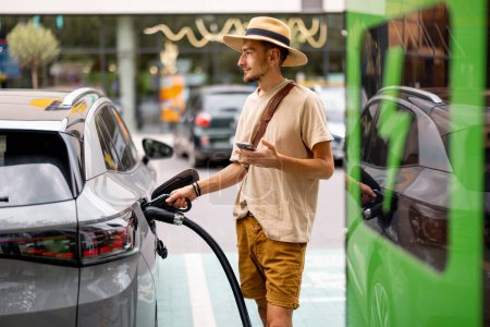 Photo for Man in hat plugs a cable in electric vehicle, while standing with phone on a public charging station outdoors. Concept of travel by electric car and green energy for driving - Royalty Free Image