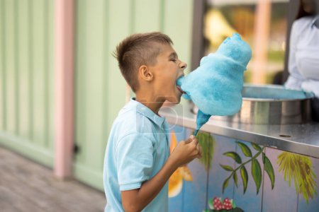 Foto de Happy boy come dulces de algodón azul mientras visita el parque de atracciones, pasa el verano felizmente, concepto de entretenimiento y vacaciones - Imagen libre de derechos