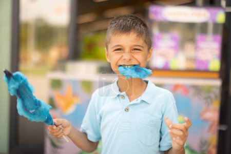 Foto de Happy boy come dulces de algodón azul mientras visita el parque de atracciones, pasa el verano felizmente, concepto de entretenimiento y vacaciones - Imagen libre de derechos