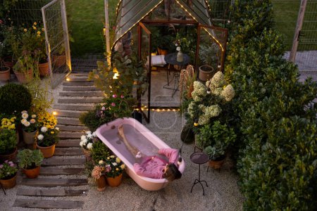 Téléchargez les photos : Cour arrière confortable avec une femme se baignant dans une baignoire rose à l'extérieur au crépuscule. Concept de détente et de beauté - en image libre de droit