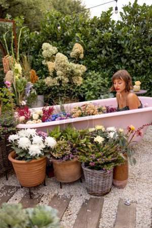 Téléchargez les photos : Femme couchée dans une baignoire rose avec beaucoup de fleurs dans le jardin à l'extérieur. Concept de beauté, thérapie bizarre et fleurie - en image libre de droit