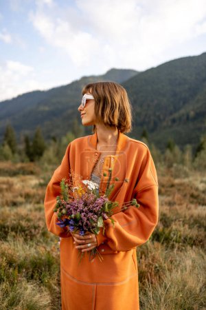 Foto de Retrato de estilo de vida de mujer en ropa naranja con ramo de campo en las montañas durante un otoño - Imagen libre de derechos