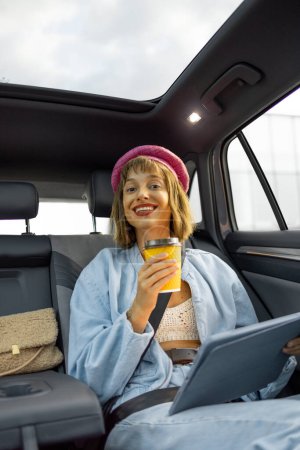 Foto de Retrato de una joven mujer alegre y elegante con tableta digital y taza de café sentado en el asiento trasero de un coche mientras conduce en la ciudad - Imagen libre de derechos