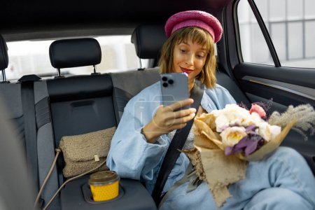 Téléchargez les photos : Jeune femme élégante utilise un téléphone intelligent tout en étant assis avec un bouquet de fleurs sur le siège arrière de la voiture sur la route. Concept de transport et mode de vie urbain - en image libre de droit