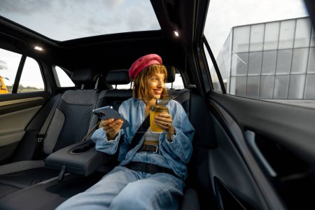 Foto de Mujer joven y elegante utiliza el teléfono inteligente mientras está sentado relajado con una taza de café en el asiento trasero del coche sobre la marcha. Amplia vista angular, coche moderno con techo panorámico - Imagen libre de derechos
