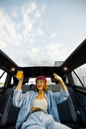 Téléchargez les photos : Jeune femme élégante aime la musique tout en voyageant en voiture, assis détendu avec une tasse de café sur le siège arrière. Vue grand angle, voiture moderne avec toit panoramique - en image libre de droit