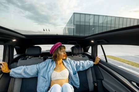 Téléchargez les photos : Jeune femme élégante assise détendue sur le siège arrière d'une voiture moderne tout en voyageant en ville. Voiture moderne avec toit panoramique - en image libre de droit