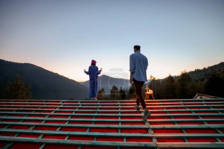 Foto de El hombre y la mujer disfrutan de una hermosa puesta de sol en las montañas, mientras están de pie en el techo de su casa inacabada. Concepto de romance, casa de ensueño y escapada a la naturaleza - Imagen libre de derechos