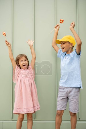 Foto de Retrato de un niño feliz con dulces caramelos en el fondo de la pared verde al aire libre, pasando el verano juntos. Niño pequeño con una chica visitando el parque de atracciones y divirtiéndose - Imagen libre de derechos