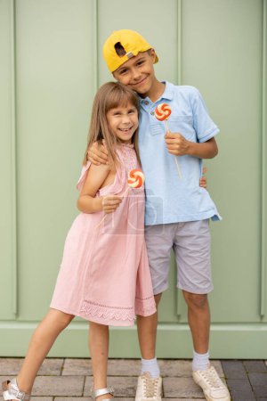 Téléchargez les photos : Portrait d'un garçon heureux avec sa petite sœur debout avec des bonbons sucrés sur fond de mur vert à l'extérieur, passer l'été ensemble. Petit garçon avec une fille visitant le parc d'attractions et s'amusant - en image libre de droit