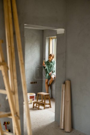 Foto de Mujer joven pinta paredes mientras hace la reparación de un nuevo apartamento. Tomando un descanso y sentado en un alféizar de la ventana. Proceso creativo del concepto de renovación y reparación del hogar - Imagen libre de derechos