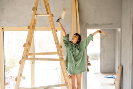Téléchargez les photos : Portrait d'une jeune femme mignonne debout joyeusement sur une échelle avec rouleau de peinture pendant le processus de réparation d'une maison. Processus créatif de rénovation et de réparation de la maison concept - en image libre de droit