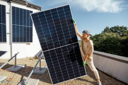 Téléchargez les photos : L'homme porte un panneau solaire lors de l'installation de la plante solaire d'un toit de sa propriété. Vue grand angle. Le concept des énergies renouvelables pour l'autoconsommation. Idée d'installation de panneaux pour les ménages - en image libre de droit