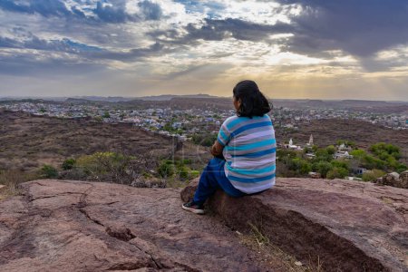 chica aislada sentada en la cima de la montaña con vista a la ciudad y el cielo naranja dramático en la imagen de la noche se toma en la India mehrangarh jodhpur rajasthan.