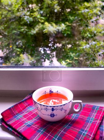 Photo for Traditional English cup of tea in a porcelain cup on a Scottish tartan cloth on a window ledge overlooking green trees. Vertical shot. - Royalty Free Image