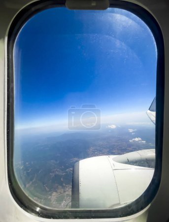 Téléchargez les photos : Cadre d'une fenêtre d'avion donnant sur le paysage montagneux et le ciel bleu, l'un des moteurs et une partie de l'aile au premier plan. Espace de copie. - en image libre de droit