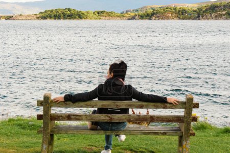 Téléchargez les photos : Plan arrière d'une jeune femme caucasienne aux cheveux bruns assise sur un banc en bois, regardant la mer détendue dans la baie de Kyleakin sur l'île de Skye. Concentration sélective sur l'arrière-plan. Plan horizontal. - en image libre de droit