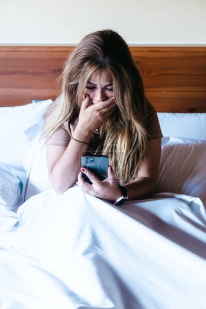 Foto de Mujer joven sentada en la cama mirando su smartphone con una expresión preocupada, conmocionada y sorprendida mientras cubría su boca con la mano. Vertical con espacio de copia. - Imagen libre de derechos