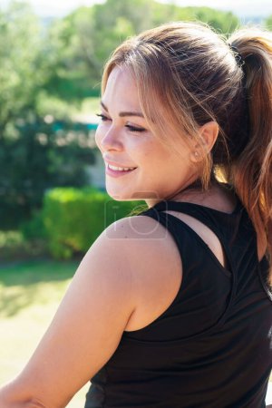 Photo for Side view of a Spanish young woman with pony tail smiling and looking away with nature background. Vertical. - Royalty Free Image