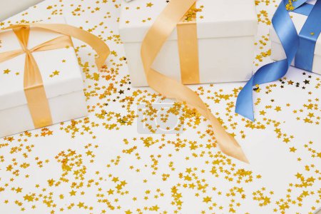 White wrapped gift boxes with gold and blue ribbons, and golden star shaped glitter on white background with copy space