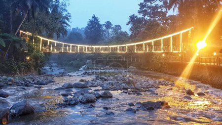 A beautifully lit suspension bridge over a rocky river at dusk, surrounded by lush greenery. Perfect for themes of travel, nature, scenic landscapes, and night photography