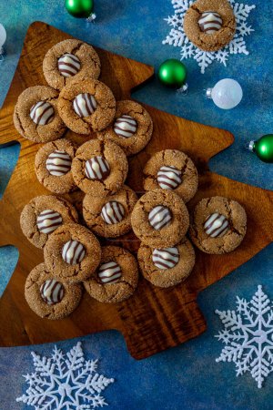 Foto de Close up of holiday cookie display of gingerbread blossom cookies sitting on wooden Christmas tree cutting board, on blue abstract background, with white snoflakes and Christmas ornaments - Imagen libre de derechos