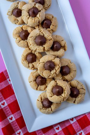 Close up of plate of peanut butter blossom cookies, stacked on white rectangle plate with heart red and white checked napkin sitting on pink background