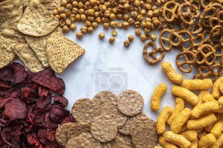 Background of assorted plant based snacks with chia quinoa torilla chips, sweet potato pretzels, chickpea cheese puffs, red beet chips and pecan nut crackers sitting on white marble counter, shot from above