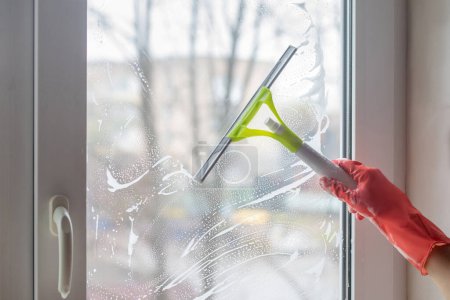 Téléchargez les photos : Le nettoyeur lave la vitre savonneuse à la main avec un grattoir. - en image libre de droit