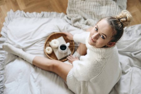 Beautiful blond caucasian girl, young woman in white knitted sweater, socks holding cup of coffee. Winter, autumn breakfast in bed. Wicker tray, white little pumpkins, blurred background, top view. Poster 620703128